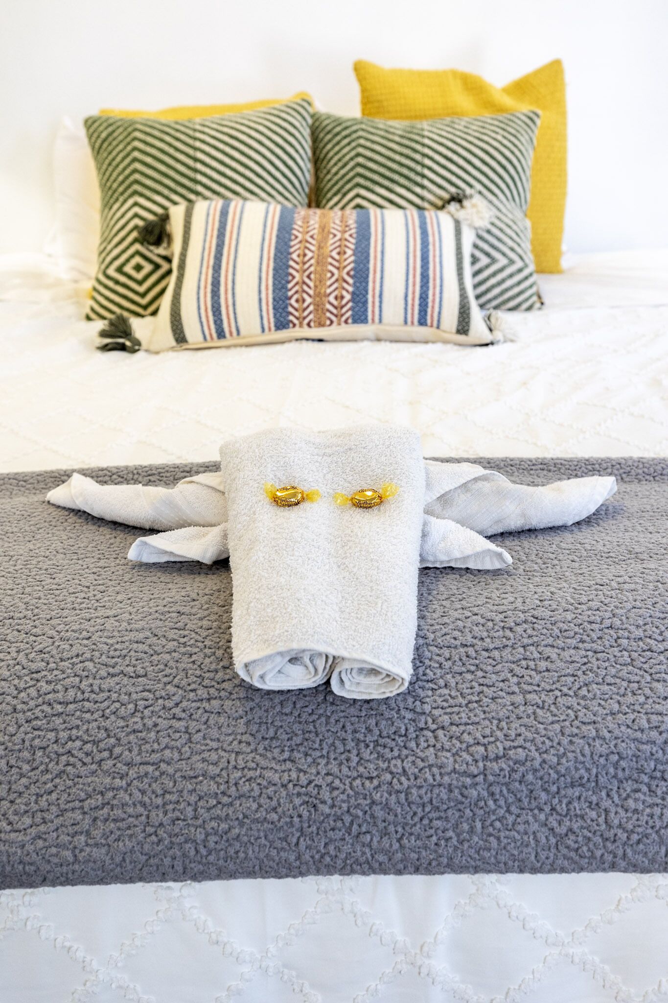 A neatly made bed with a grey textured blanket, a white towel with a gold brooch, and a variety of patterned pillows in green, yellow, and multicolored stripes.