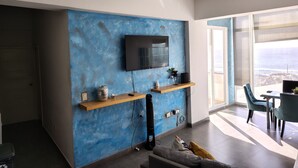 Un salon moderne avec un mur bleu à texture, un téléviseur fixé au mur, une étagère en bois ornée d’objets décoratifs, un canapé et un coin repas avec des chaises.