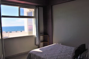 Une chambre avec un lit, une fenêtre donnant sur la mer et une petite étagère.