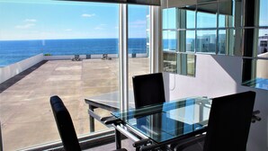 Une pièce moderne avec une table et des chaises en verre, de grandes fenêtres donnant sur l’océan et un balcon avec des sièges d’extérieur.