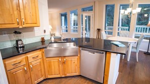 Una cocina moderna con armarios de madera, un lavamanos de acero inoxidable y un lavavajillas. Hay un comedor con una mesa y sillas blancas, y ventanas amplias que ofrecen vistas al exterior.