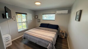 A bedroom with a bed, two windows, an air conditioner, a small bedside table, and a wall-mounted TV. - Perfect slice of peace and quiet by<br> (Lake Placid)