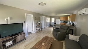 A spacious living room with a flat-screen TV, a gray sofa, and a coffee table. There is a kitchen area with wooden cabinets and a refrigerator. - Perfect slice of peace and quiet by<br> (Lake Placid)