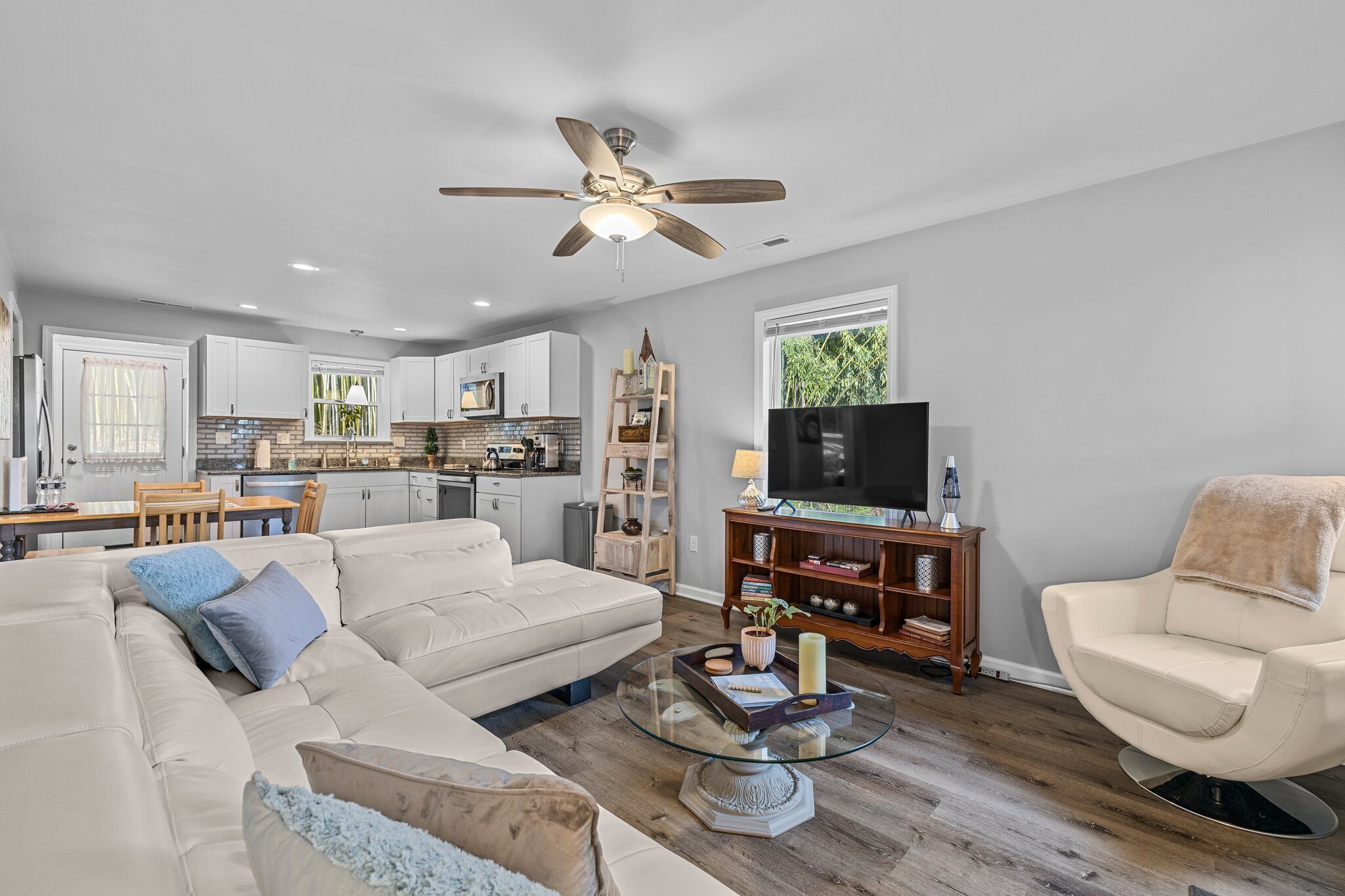 A modern living room with a sofa, coffee table, and a TV. There is a kitchen in the background with cabinets and a dining area.