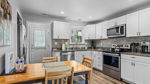 A modern kitchen with white cabinets, a granite countertop, stainless steel appliances, and a dining area with a wooden table and chairs. - 1/4 Mile to Ecusta Trail • Firepit • 1G Fiber Wi-Fi • Bamboo Bungalow (Hendersonville)