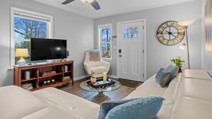 A living room with a white sofa, a glass coffee table, a television, a bookshelf, a ceiling fan, and a clock on the wall. - 1/4 Mile to Ecusta Trail • Firepit • 1G Fiber Wi-Fi • Bamboo Bungalow (Hendersonville)