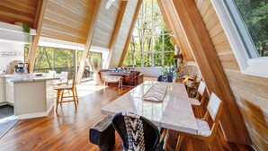 A spacious loft-style room with a high ceiling, wooden beams, a large dining table, and a cozy seating area. - A-Frame of Mind  •  Fenced Yard -  Lake Access - AC - Lake View - One-of-a-Kind (Lake Arrowhead)