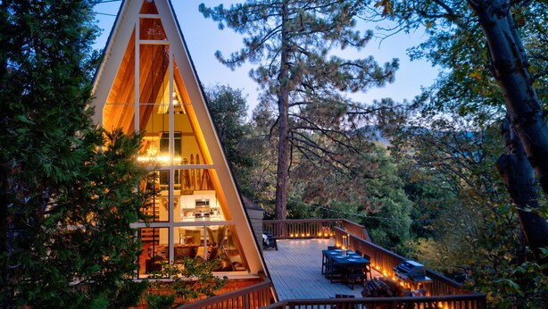 A modern, triangular-shaped cabin with a deck and outdoor seating area, surrounded by trees. - A-Frame of Mind  •  Fenced Yard -  Lake Access - AC - Lake View - One-of-a-Kind (Lake Arrowhead)