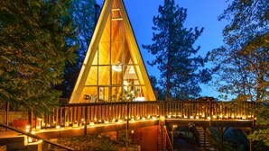 A triangular glass-roofed building with a wooden deck and railing, surrounded by trees. - A-Frame of Mind  •  Fenced Yard -  Lake Access - AC - Lake View - One-of-a-Kind (Lake Arrowhead)