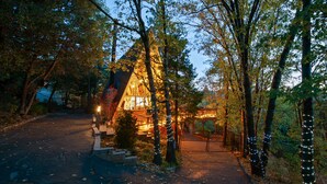 A rustic cabin with a sloped roof, surrounded by trees and illuminated by string lights. - A-Frame of Mind  •  Fenced Yard -  Lake Access - AC - Lake View - One-of-a-Kind (Lake Arrowhead)