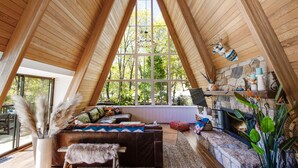 A cozy room with a sloped ceiling, a stone fireplace, a comfortable seating area, and large windows offering a view of the outdoors. - A-Frame of Mind  •  Fenced Yard -  Lake Access - AC - Lake View - One-of-a-Kind (Lake Arrowhead)