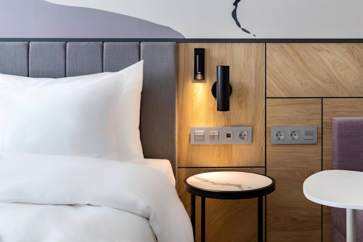 Une chambre d’hôtel moderne équipée d’un lit, d’une petite table ronde, d’une lampe fixée au mur et de prises électriques.