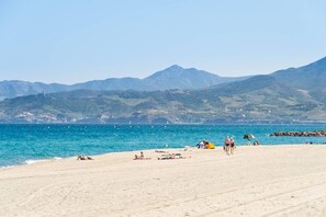Una playa con gente tomando el sol y paseando, cielo despejado y montañas al fondo.