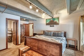 Un dormitorio con cama de madera, una mesita de noche pequeña, un banco, un cuadro en la pared y una puerta que da a un baño con toallero.