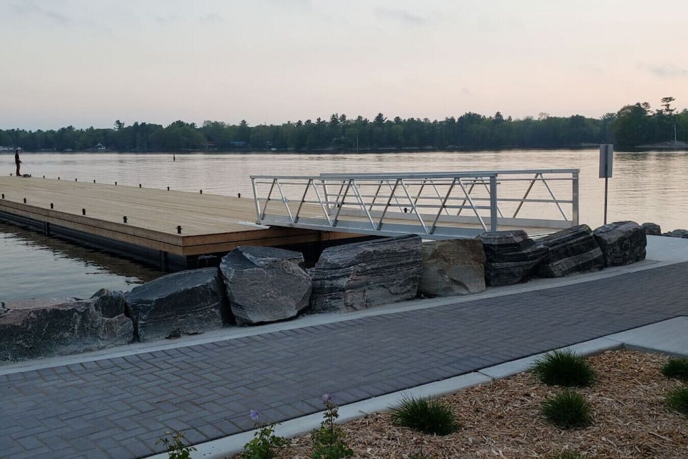 Eine Holzstele, die sich in ruhiges Wasser erstreckt, mit Metallgeländer und großen Felsen am Ufer.