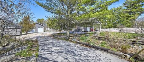 A paved pathway leading to a building with a covered porch area, surrounded by trees and a gravel area.