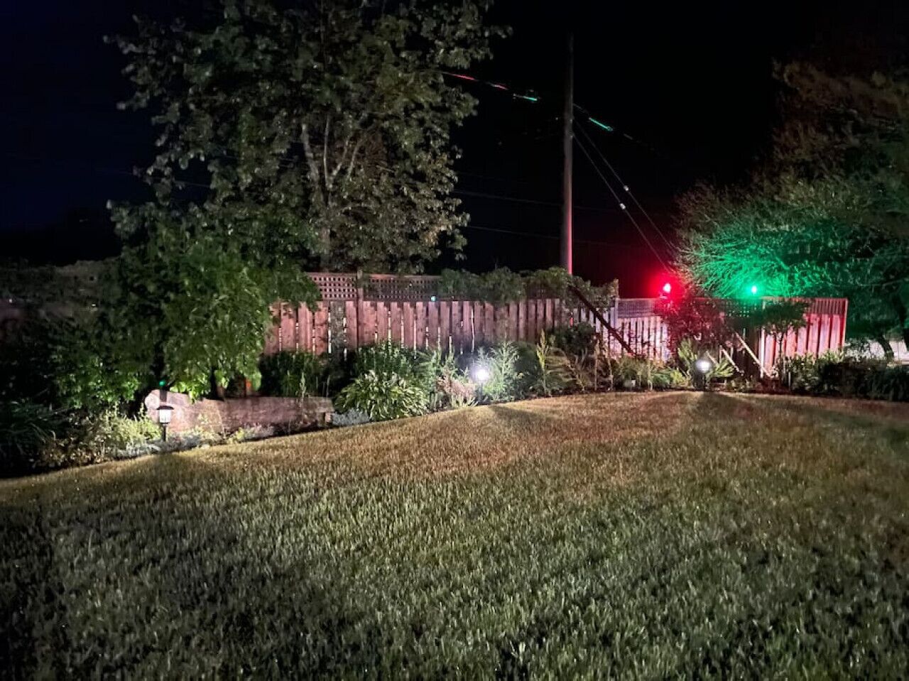 A nighttime view of a residential area with a well-lit house, a red fence, and a green-lit streetlight.