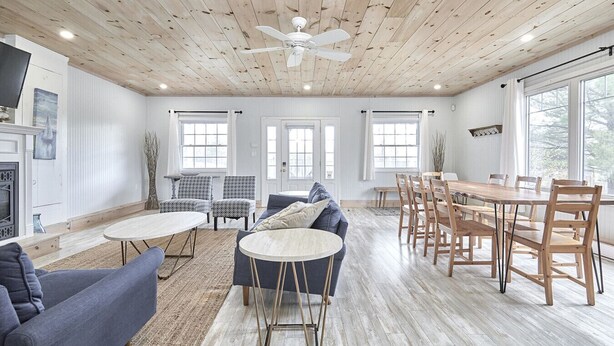 A spacious living room with a wooden ceiling, a fireplace, a dining area with wooden chairs, and large windows.
