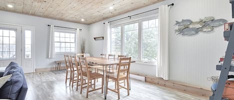 A spacious dining area with a wooden table and chairs, large windows, and a view of trees outside.