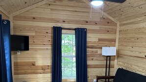 Habitación con paredes y techo de madera, ventilador de techo, ventana con cortinas, sofá negro, sillón blanco y televisor de pantalla plana montado en la pared.