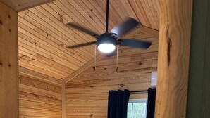 Un dormitorio de cabaña de madera con una cama, un ventilador de techo, una ventana con cortinas y una mesita de noche con lámpara.