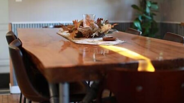Une table à manger en bois, entourée de chaises, agrémentée d’un centre de table composé de feuilles séchées et d’un tableau encadré accroché au mur.