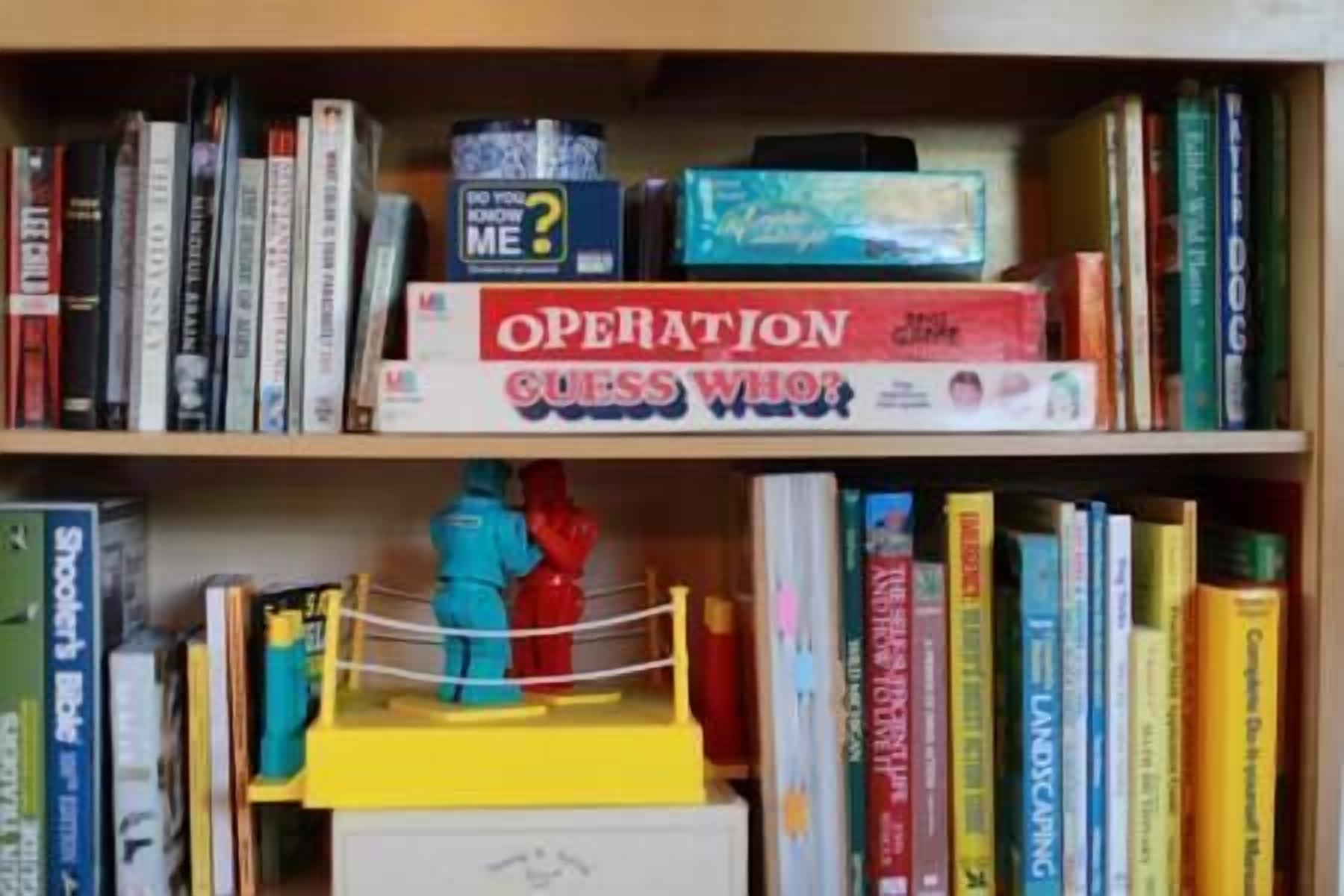 A bookshelf with various books and two games, including 'Operation' and 'Guess Who?'.