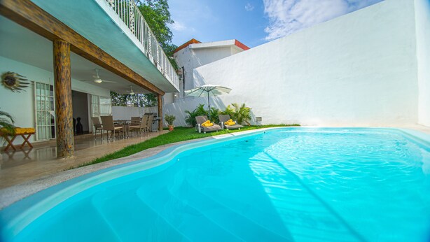 A swimming pool with lounge chairs and a table, surrounded by a white wall and greenery.