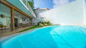 Una piscina con sillones de descanso y una mesa, rodeada por un muro blanco y vegetación.