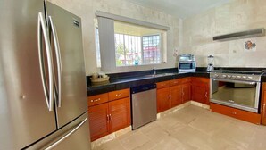 A kitchen with a stainless steel refrigerator, a microwave, an oven, wooden cabinets, and a window with bars.