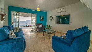A living room with two blue sofas, a glass coffee table, and a grey armchair. There is a ceiling fan, an air conditioning unit, and a large window with a view. - Beautiful home with private pool (Puerto Vallarta)