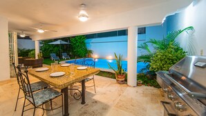 An outdoor patio with a dining table set for four, surrounded by chairs, a grill, and a pool in the background. - Beautiful home with private pool (Puerto Vallarta)