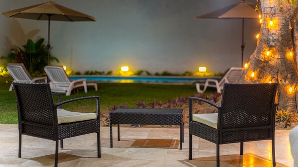 An outdoor seating area with wicker chairs and a table, a pool in the background, and string lights on a tree. - Spacious home with private heated pool (Puerto Vallarta)