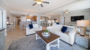 A spacious living room with a white sofa, a wooden coffee table, and a chandelier. There is a ceiling fan and a TV mounted on the wall. - Azure 615 ~ 4BR Oceanfront Condo ~ Sleeps 10 ~ Spectacular Penthouse Views! (Fort Walton Beach)