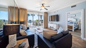 Ein modernes Wohnzimmer mit einer Couch, einem Couchtisch und einem Fernseher. Es gibt einen Balkon mit Vorhängen und einem Deckenventilator.