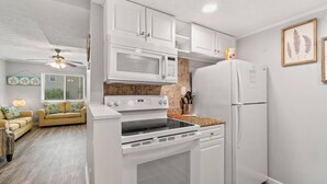 A modern kitchen with white cabinetry, a microwave, stove, oven, and refrigerator. There is a window with blinds and a ceiling fan. - Misty Harbor Townhome #15 (Panama City Beach)
