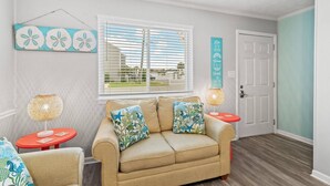 A living room with a beige sofa, two side tables, a window with blinds, and a door. - Misty Harbor Townhome #15 (Panama City Beach)