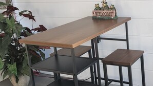 Een houten tafel met een 'Welcome' bord en kikkerbeeldjes, voor een witte muur met een 'Cute & Cozy' bord.