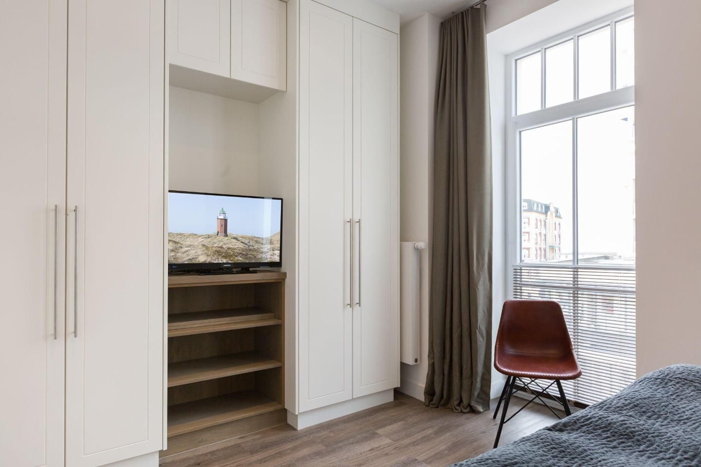 A room with a TV on a wooden shelf, a red chair, and a window with curtains.