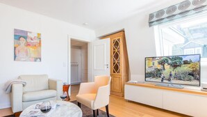Un salon moderne avec un canapé, un fauteuil et une table basse. Un téléviseur est fixé au mur, et une porte donne accès à une autre pièce.