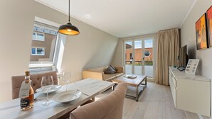 Habitación de hotel moderna con zona de comedor, sofá, televisor y balcón con vistas a la ciudad.