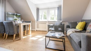 A modern living room with a dining area, a grey sofa with a yellow pillow, and a table with a magazine and wine glasses. - Komfortabel. Ruhig. Gemütlich. - Ringhoog Hüs Whg. 3 "wiesenblick" - Objekt-id: Rhh/03 - Ringhoog (Sylt)