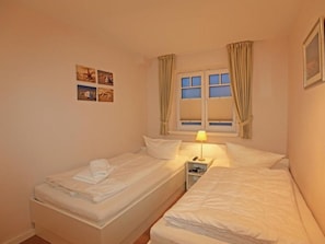 Une chambre avec deux lits, une table de chevet avec une lampe, une fenêtre avec des rideaux et un mur orné de quatre petits tableaux.
