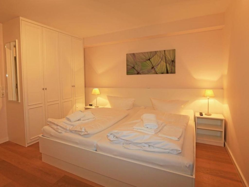 A double bed with white linens, two bedside tables with lamps, a wardrobe, and a wall-mounted abstract painting.