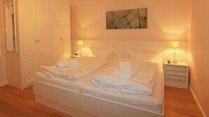 Un lit double avec du linge de lit blanc, deux tables de chevet avec des lampes, une armoire et un tableau abstrait accroché au mur.