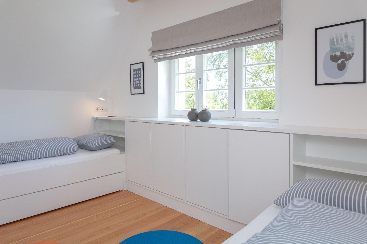 A modern, minimalist bedroom with a built-in bed, a window with a view of greenery, and a small seating area with cushions.