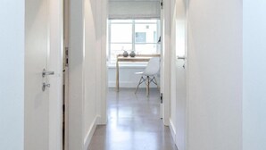 A narrow hallway with white walls and a wooden ceiling beam. A door is visible on the left, and a window with a table and chair is seen in the distance. - House Mühlenweg, Germany (Sylt)