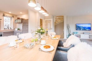 Un espace repas moderne avec une table en bois dressée pour le repas, une cuisine en arrière-plan et un téléviseur fixé au mur.