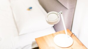 Une lampe blanche posée sur une table d’appoint en bois, un coussin blanc et un autre coussin blanc sur un canapé gris.