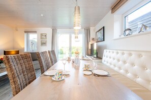 A dining area with a long table, wicker chairs, a white cushioned bench, and a hanging light fixture. - Haus Waldwinkel 18, Whg. Auszeit - Exklusiv. Strandnah. Auszeit. - Haus Waldwinkel 18, Whg (Sylt)